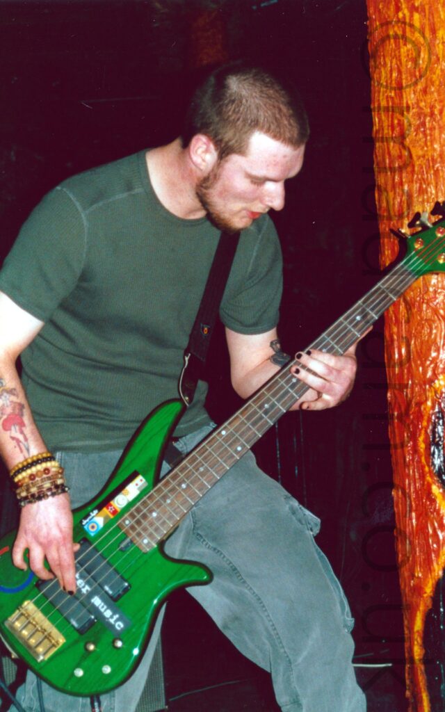 A man with short dark hair and beard, wearing a green t-shirt and a grey pair of cargo pants, playing a bright green 5-stringed bass guitar. His right wrist is covered in an array of beaded bracelet, while his right forearm has a tattoo of a red love heart with a large knife through it. His left forearm has a black tattoo which is partially hidden behind the neck of the bass. He is slightly hunched over, looking down at his left hand. In the background, an orange pole fills the right of the frame, glistening wetly, with a visceral, organic appearance, with black walls and ceiling filling the rest of the frame.