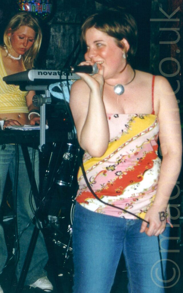 A woman with brown hair that has been cut in a bob, wearing a scrappy top with a series of diagonal coloured stripes on the front, and a blue pair of jeans, singing into a microphone held in her right hand, with her head turned to look off to her right. In the background, on the left of the frame, is a woman with blonde hair in a long bob, wearing a yellow top and blue jeans, standing behind a rack of keyboards, her hands resting on the bottom board. Black walls fill the rest of the background.