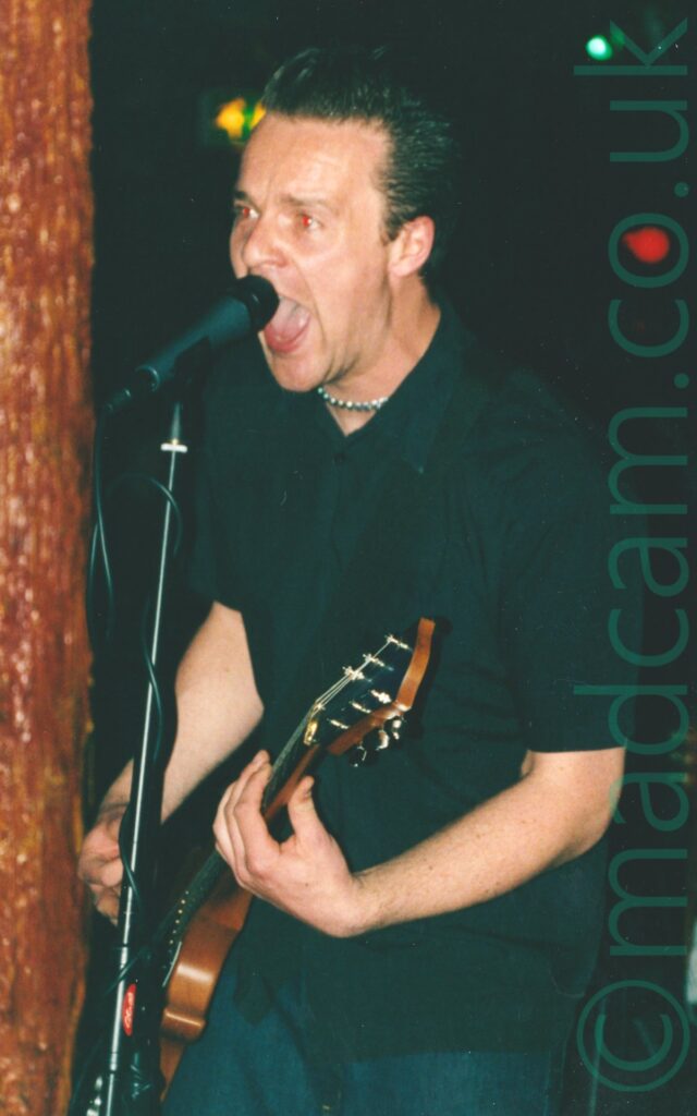 A man with slicked-back short hair on a stage, wearing a black shirt and a blue pair of jeans, playing a black and brown electric guitar, and shouting into a black microphone on a stand in front of him. An amber-coloured pole is in the background behind him, looking rather viscous and organic. Black walls fill the background, with spots of yellow, blue, and red lights on the wall.