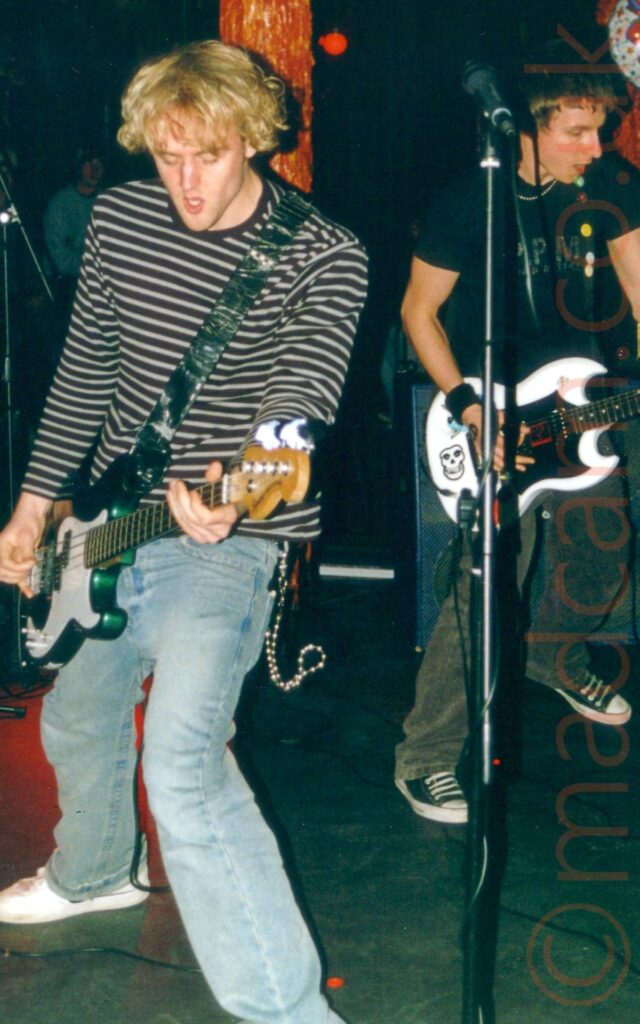 Two men performing on a stage as part of a band. On the left is a man with floppy blond hair, wearing a long sleeved top with thin horizontal black and white stripes, and a faded pair of blue jeans, playing a green and white bass guitar. On the right is a man with short dark hair, wearing a black t-shirt with grey text on the front and black jeans, playing a white and black electric guitar. A white foil ballon with colourful text "Happy 21st Birthday" hangs in the air behind his head, looking for all the world like a comic book speech bubble. There are a pair of amber poles in the background, that have been moulded and painted to resemble something moist, viscous, and organic. People can be seen in the darkness in the background.