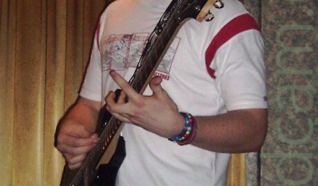 A man with short, dark, spiky hair, wearing a white t-shirt with red bands around the shoulders, and grey and red squares on the chest, as well as blue jeans, playing a black electric guitar while looking down at his left hand. Golden velvet curtains cover the wall on the left of the background, with stippled green and red wallpaper on the right.