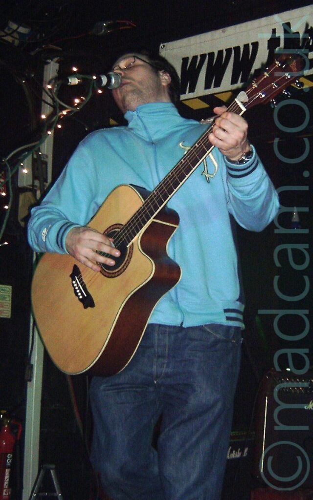 A man with short dark hair, wearing a blue light jacket zipped up to his neck and a blue pair of jeans, playing a brown acoustic guitar, and leaning backwards while singing into a microphone on a stand. White fairy lights are wrapped around the mic stand. Black walls and ceiling fill the background.