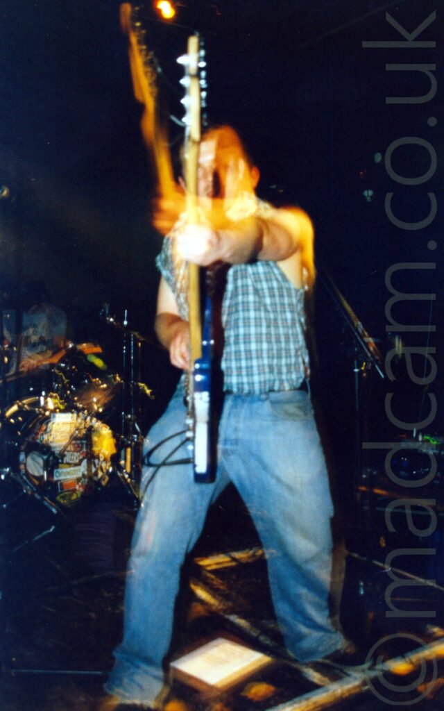 A man with short dark hair, wearing a checked shirt with the sleeves removed and a blue pair of jeans, playing a blue bass guitar, with the base of the guitar against his leg, and the head pointed towards the camera. A drum kit is visible in the background, on the left. A long exposure and his movements result in the stage lights creating secondary images in golden light contrasting with the black walls and ceiling in the background.