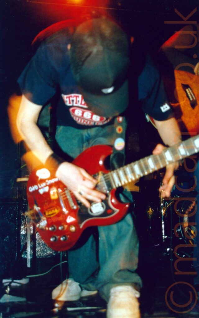 A man wearing a black baseball cap and a dark blue t-shirt with an indistinct red and white logo across the chest, and a blue pair of jeans, crouched over and playing a red electric guitar held almost at ground level. A red light shines above his head, while golden light glows around his left arm. Black walls and ceiling fill the rest of the background.