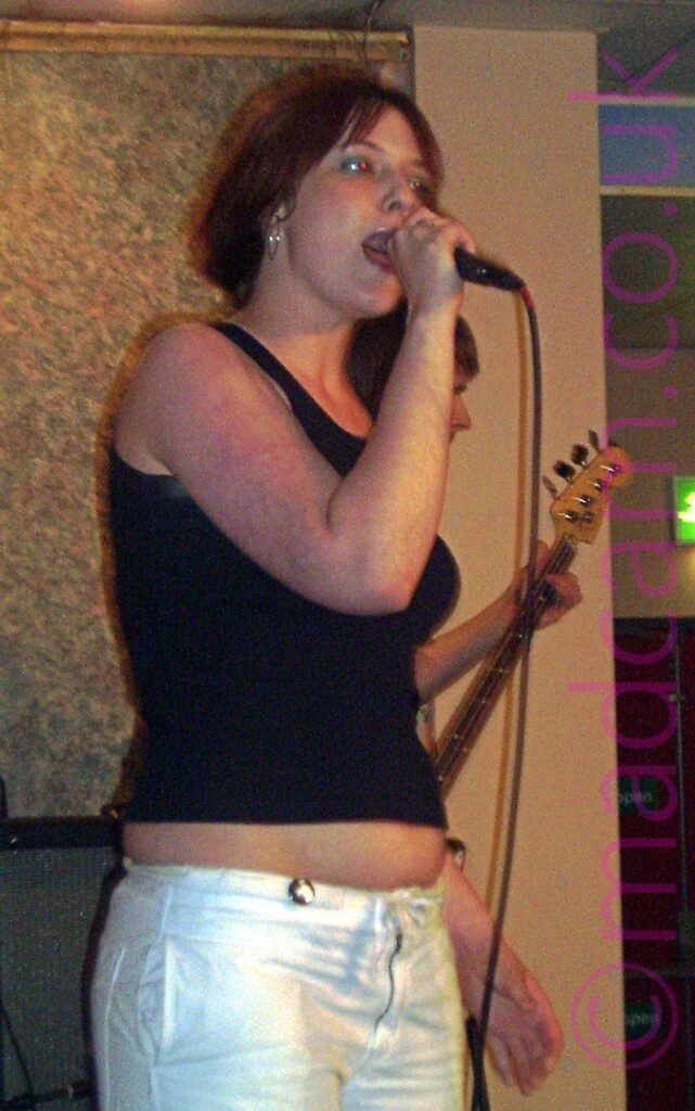 A woman with red hair, wearing a black tank top and white trousers, singing into a microphone held in her right hand, as she stares off to the right of the camera. A man can be seen behind her, playing a bass guitar, in front of a yellow-painted concrete support post. A rather muted but colourful wallpaper covers the wall on the left of the frame, with white ceiling panels at the top.
