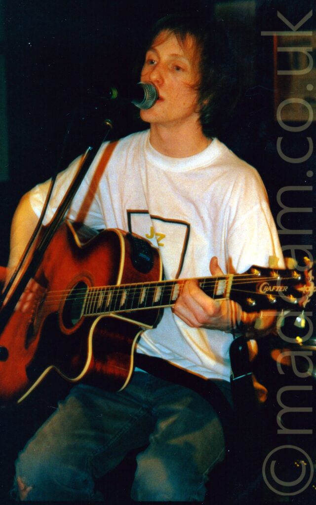 A man with floppy dark hair, wearing a white t-shirt with the black outline of a square and the gold letters "J Z" in the middle, and playing a brown acoustic guitar. There is a microphone on a stand in front of him, which he is singing into, while sitting on a stool. Black walls fill the ceiling.