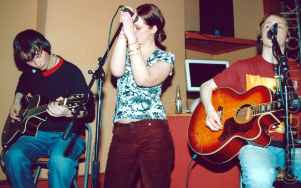 Three people performing on a stage as part of a band. On the left is a man with mid-length dark hair, wearing a dark blue jumper over a red t-shirt and blue jeans, playing a dark brown acoustic guitar while sitting on a stool, looking down at his left hand. In the middle is a woman with her brown hair tied back, wearing a white top with a green leafy print all over, and mauve jeans, using both hands to hold on to a microphone mounted on a stand in front of her, while the looks down and to her right. On the right is a man with short but wavy brown hair, wearing a mauve t-shirt with a large yellow square on the front, playing a brown acoustic guitar while singing into a microphone on a stand in front of his mouth. Beige walls with red shelves fill the background.