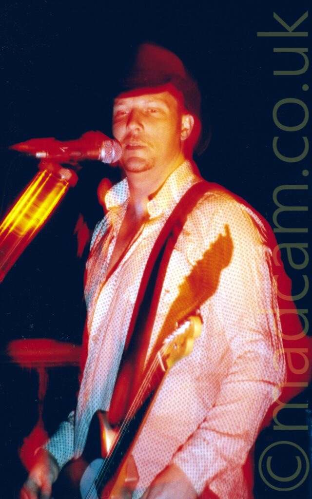 A man standing by a microphone on a stand, looking out over the audience. He is wearing a dark stetson, and a white shirt covered in small dark dots. A grey electric guitar hangs by a strap over his left shoulder. Black walls and ceiling fill the background, while a long exposure and red and yellow stage lights creates red and yellow auras around him and the microphone.