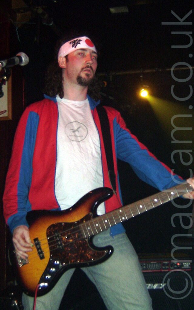 A man with long dark hair tied back, and a brown goatee and moustache, wearing a red and blight light jacket over a white t-shirt, and slightly grubby blue jeans. He is playing a brown and amber bass guitar, and giving the camera some serious side-eye. A white headband with a red circle on the front and some Japanese writing on the side is wrapped around his head, tying his hair back. A single yellow light shines out from a lighting rig in the black background.