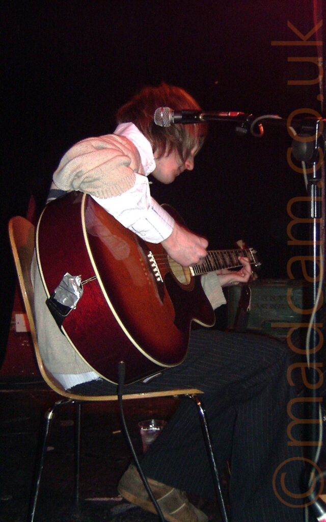 A man with shaggy blond hair, wearing a cream jumper over a white shirt, and black trousers with a white pinstripe pattern, sitting on a chair facing to the right and playing a brown acoustic guitar. A red stage light is coming from the top left corner of the frame, with black walls and ceiling filling the rest of the frame.