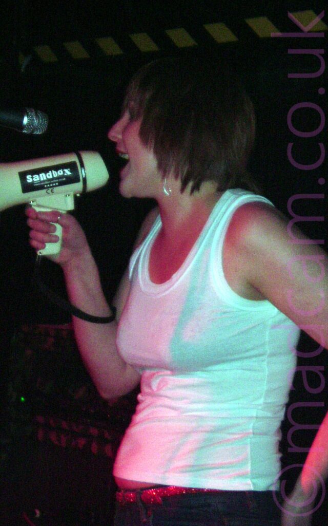 A woman with chin length dark hair, wearing a white tank top over blue jeans, singing into a white megaphone held in her right hand pointed off to the side of a microphone in front of her. A black label with white text "Sandbox" is on the side of the megaphone. Black walls and ceiling fill the background, with yellow and black hazard tape on the wall behind her.