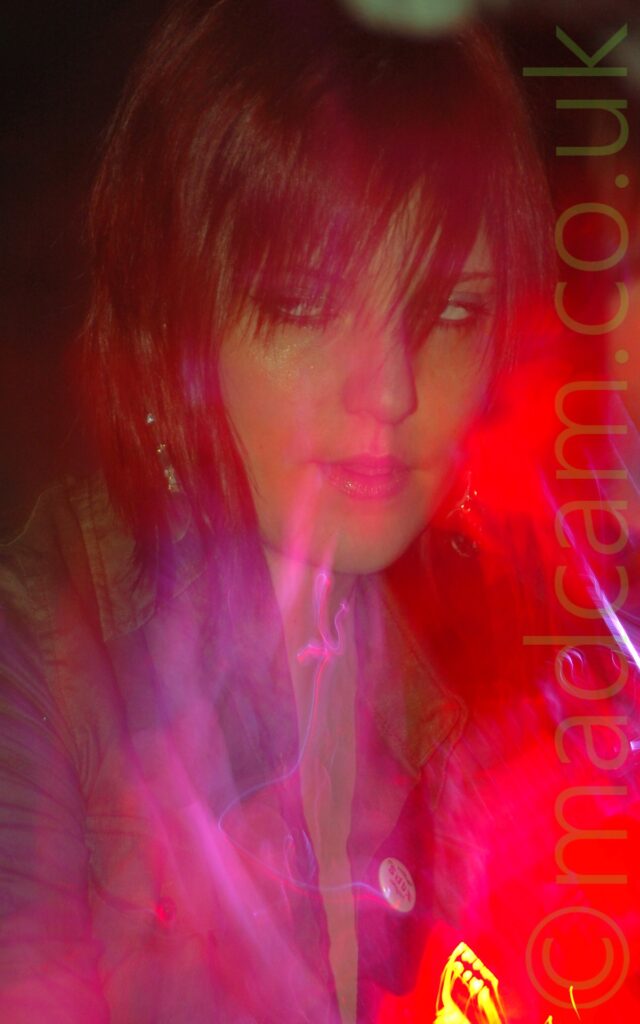 A woman with dark red hair, wearing a satiny green shirt, staring moodily off to her right through her fringe. Red light floods the right of the frame, with purple auras and streaks of light hinting at a snap zoom out, with suggestions of a zoomed-out shot of here holding a guitar. Black walls fill the background.