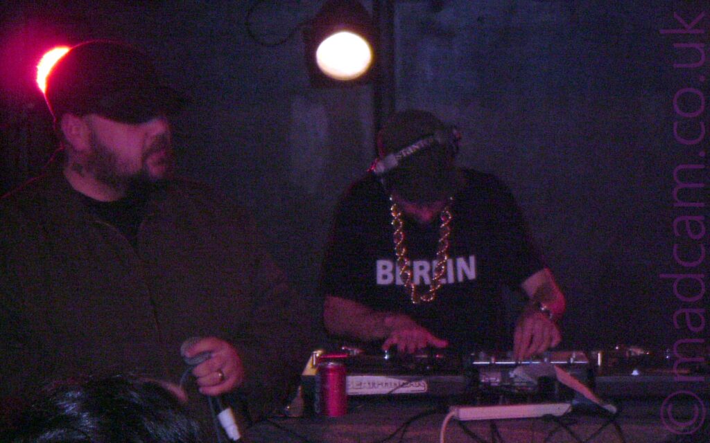 Slightly grainy shot of 2 men performing on a stage. On the left is a man with a short, dark beard, wearing a black baseball cap and a dark grey jacket over a black t-shirt, holding a microphone in his left hand. A red stagelight can be seen shining behind his head. On the right is a man with short dark hair, wearing a pair of chunky headphones on his head, and a black t-shirt with the word "Berlin" in white on the front, and with a chunky gold rope chain around his neck. He has a pair of turntables in front of him, and is manipulating one of the controls on a mixer with his left hand while scratching on one of the turntables with his right. A white stagelight shines down over his right shoulder. Black walls fill the background.