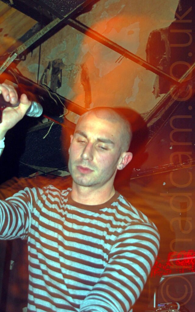 Low-down shot of a man with a shaven head, wearing a grey and brown banded t-shirt, and holding a microphone on a stand in his right hand. A rather patchy ceiling, with brown and black panels, fills the background behind him.