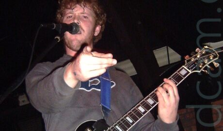 A man with short, brown, curly hair and a short, straggly beard, wearing a grey hoody over a grey t-shirt and a blue pair of jeans. He is holding a black electric guitar with his left hand, while pointing at some idiot with a camera with his right hand. There are several white logos on the guitar - one reads "Biffy Clyro", another reads "Gotta Love Hotties". Black walls and ceilings with several stage lights fill the rest of the frame.