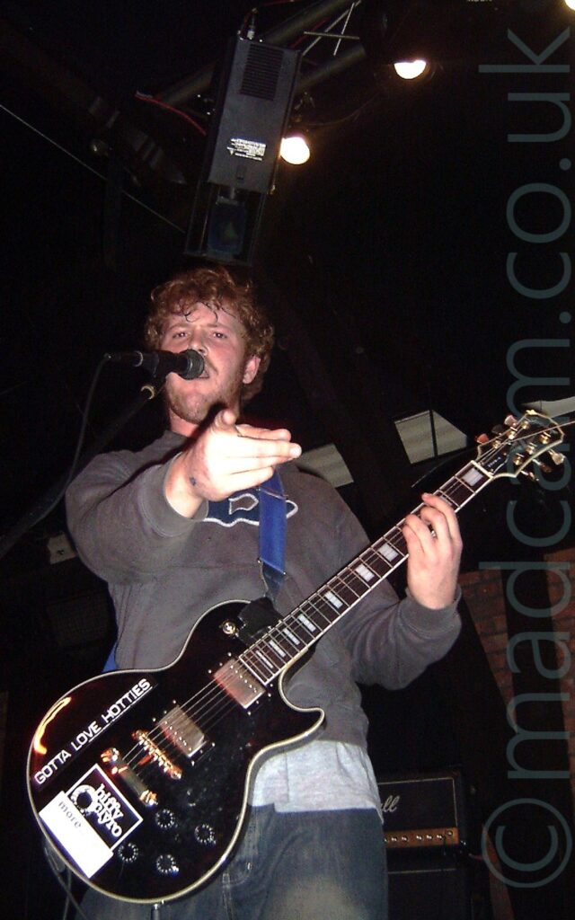 A man with short, brown, curly hair and a short, straggly beard, wearing a grey hoody over a grey t-shirt and a blue pair of jeans. He is holding a black electric guitar with his left hand, while pointing at some idiot with a camera with his right hand. There are several white logos on the guitar - one reads "Biffy Clyro", another reads "Gotta Love Hotties". Black walls and ceilings with several stage lights fill the rest of the frame.
