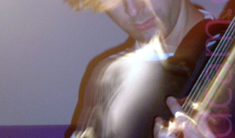 A man with floppy brown hair, wearing a black top with white piping around the neck and sleeves, and playing a bass guitar, while looking down at his left hand. The wall in the background is grey, with a purple and red stripe running across the middle.