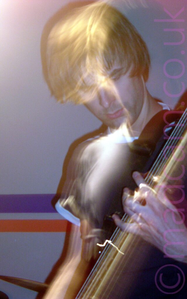 A man with floppy brown hair, wearing a black top with white piping around the neck and sleeves, and playing a bass guitar, while looking down at his left hand. The wall in the background is grey, with a purple and red stripe running across the middle.