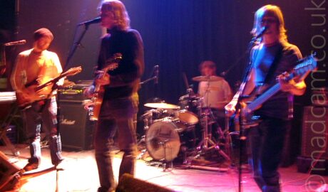 4 men performing on a stage as part of a band. On the left is a man wearing a woollen hat, and wearing a long sleeved white top, with blue jeans, and playing a black and white electric guitar. In the middle is a man with wavy blond hair, wearing a dark rollneck pullover and jeans, playing an electric guitar, and singing into a microphone on a stand in front of him. At the back of the stage, a man with curly dark hair and wearing a white t-shirt is sitting behind a black drumkit, playing a riff with his right hand. On the right is a man with long blond hair, wearing a bluet-shirt and jeans, playing a black bass guitar while singing into a microphone on a stand. A long exposure without flash shows movement by the band, with small secondary images and yellow and blue auras from the stagelights around some parts of their bodies and instruments.
