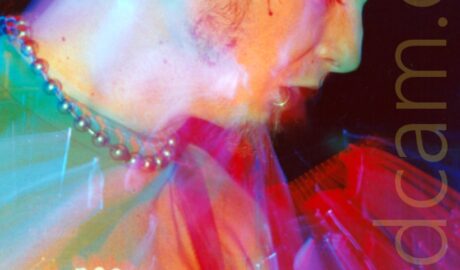 Long exposure snap zoom close-up of the face of a man facing to the right, with dark hair that is very short on the sides and fairly long on top, wearing a green t-shirt, and looking down and to the right of the frame. A flash has frozen him in place, and a long exposure mixed with a snap zoom out gives a sense of movement, with added red, yellow, green and blue stage lights going off, painting the frame with coloured lights.