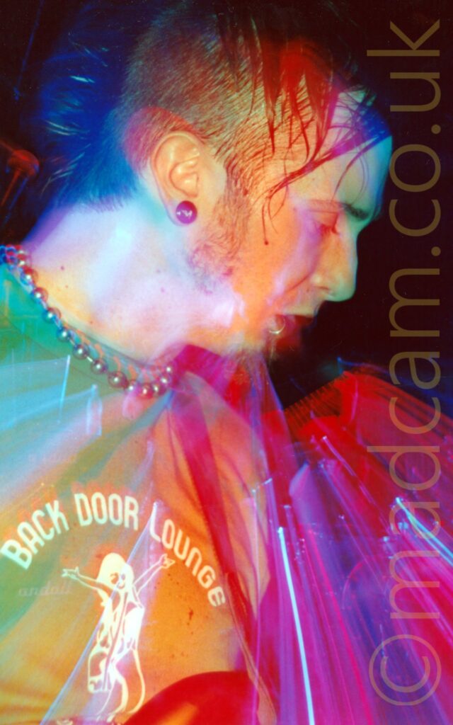 Long exposure snap zoom close-up of the face of a man facing to the right, with dark hair that is very short on the sides and fairly long on top, wearing a green t-shirt, and looking down and to the right of the frame. A flash has frozen him in place, and a long exposure mixed with a snap zoom out gives a sense of movement, with added red, yellow, green and blue stage lights going off, painting the frame with coloured lights.