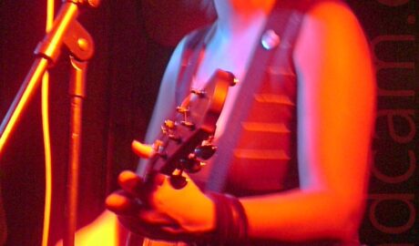 A woman with dark, chin-length hair cut in a shaggy bob, wearing a brown tank-top and denim skirt, playing an acoustic guitar, and singing into a microphone on a stand in front of her. A long exposure has been used, her right hand a blur as she strums her guitar, while the stage light cast an orange hue over everything. Dark walls and ceiling fill the background.