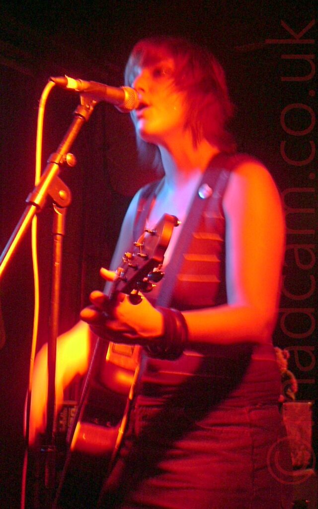 A woman with dark, chin-length hair cut in a shaggy bob, wearing a brown tank-top and denim skirt, playing an acoustic guitar, and singing into a microphone on a stand in front of her. A long exposure has been used, her right hand a blur as she strums her guitar, while the stage light cast an orange hue over everything. Dark walls and ceiling fill the background.