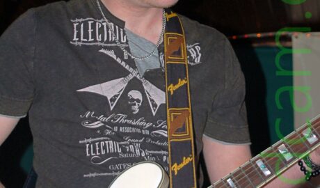 A man wearing a black knitted hat, a black t-shirt with a white print made up of mostly indecipherable text, and a blue pair of jeans. He is playing a white electric guitar, which has a black strap with yellow "Fender" text and a brown representation of a guitar head slung over his left shoulder. A microphone on a stand is in front of his head up in the top left corner. Gren walls and a brown wooden ceiling fill the background.