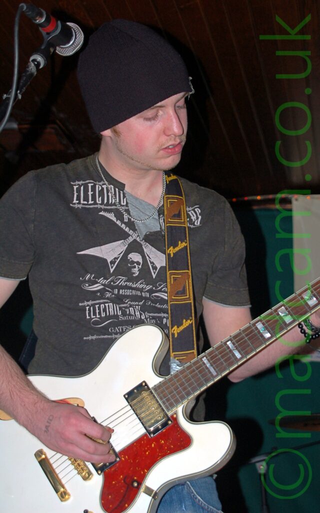 A man wearing a black knitted hat, a black t-shirt with a white print made up of mostly indecipherable text, and a blue pair of jeans. He is playing a white electric guitar, which has a black strap with yellow "Fender" text and a brown representation of a guitar head slung over his left shoulder. A microphone on a stand is in front of his head up in the top left corner. Gren walls and a brown wooden ceiling fill the background.