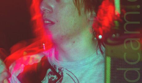 Close-up of the head and upper body of a man holding a pair of drumsticks in the air. He has straggly mid-length hair, and is wearing a grey t-shirt with an indistinct black logo across the chest. A long exposure has given his face a green cast from some of the stagelights, with red halos of light around his head, and purple blooms of light around his right wrist and sternum. Black walls flooded with red light fill the background.