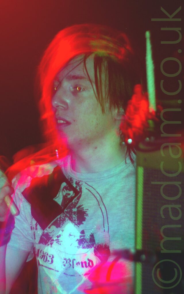 Close-up of the head and upper body of a man holding a pair of drumsticks in the air. He has straggly mid-length hair, and is wearing a grey t-shirt with an indistinct black logo across the chest. A long exposure has given his face a green cast from some of the stagelights, with red halos of light around his head, and purple blooms of light around his right wrist and sternum. Black walls flooded with red light fill the background.