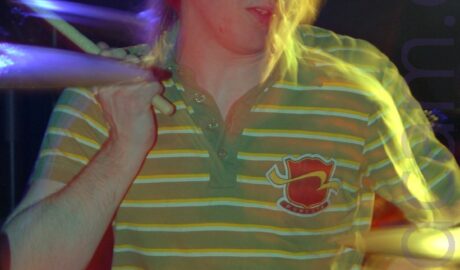 A man with floppy brown hair and wearing a green polo shirt with thin yellow and white bands around the chest and a red shield logo on the left side of the chest, sitting behind a black drum kit and waving a pair of drumsticks in the air. He has been frozen in place by a flash, but a long exposure has captured his movement afterwards, created secondary images, some of which are gold or purple, with red highlights on the drums. Black walls and ceiling fill the background.