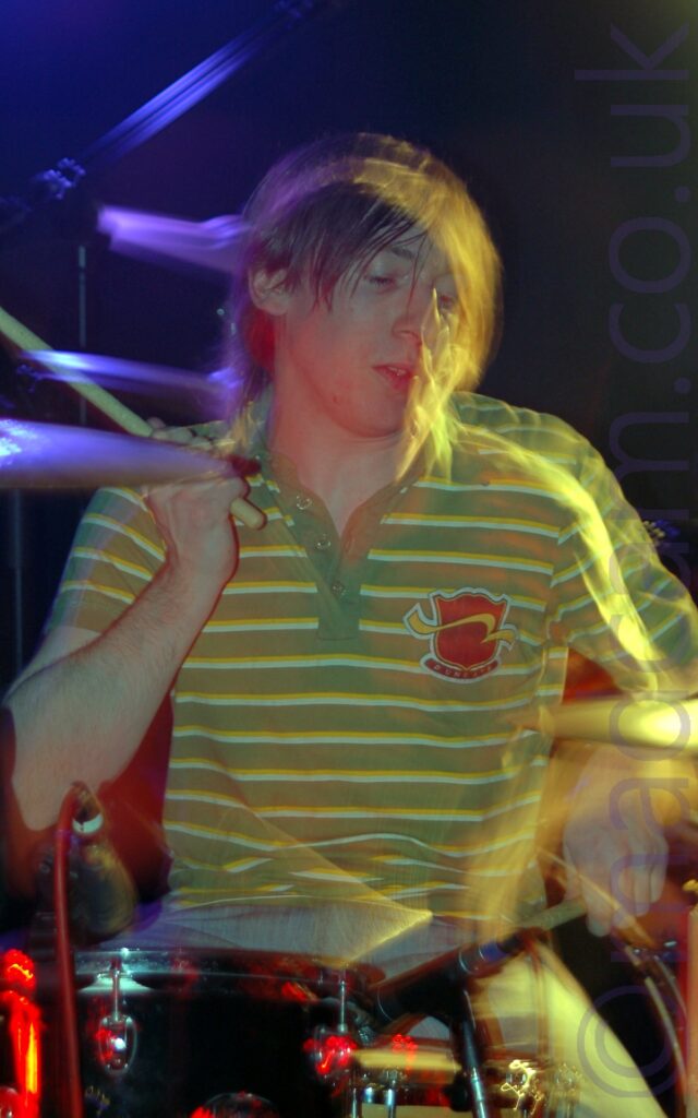 A man with floppy brown hair and wearing a green polo shirt with thin yellow and white bands around the chest and a red shield logo on the left side of the chest, sitting behind a black drum kit and waving a pair of drumsticks in the air. He has been frozen in place by a flash, but a long exposure has captured his movement afterwards, created secondary images, some of which are gold or purple, with red highlights on the drums. Black walls and ceiling fill the background.
