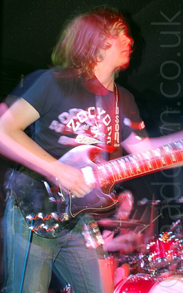 A man on a stage, performing as part of a band. He has shoulder-length, floppy brown hair, and wearing a dark blue t-shirt with whie "ZooYork" text across the front,and a blue pair of jeans. He is playing a black electric guitar attached to a black strap with white "Gibson USA" text on the front that is slung over his left shoulder. A man can be seen sitting behind a red drumkit in the bottom right corner of the frame in the background. A long exposure combine with stage lights and his movements to create secondary images and auras around him. Black wills fill the rest of the frame.