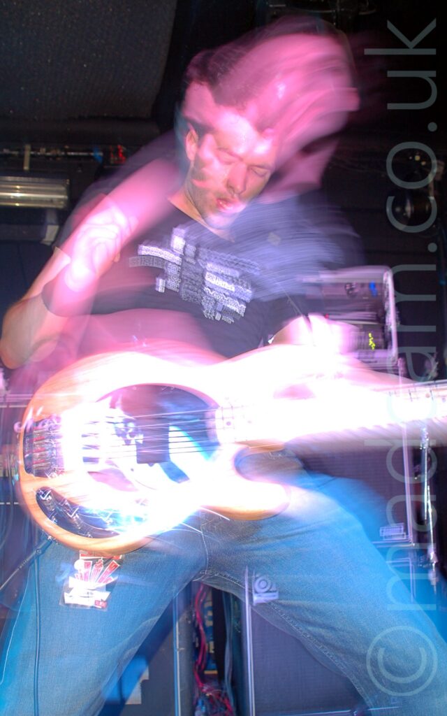 A man on stage, with short dark hair, wearing a black t-shirt with an indistinct white logo and a blue pair of jeans, playing a white bass guitar. A camera flash has frozen him in place, while a long exposure combined either flashing stage lights and his movement have created purple secondary images around his head and torso. Racks of amplifiers and speakers can be seen in the background, against black walls.