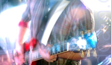 A man with short dark hair and beard, wearing a white shirt with broad red and blue vertical stripes over a grey t-shirt, and a blue pair of jeans, playing a red electric guitar, and screaming into a microphone on a stand in front of him. A long exposure, combined with his movement and the stage lights has resulted in some blurring of the image, with explosions of white light around him, reflecting off his guitar and face.