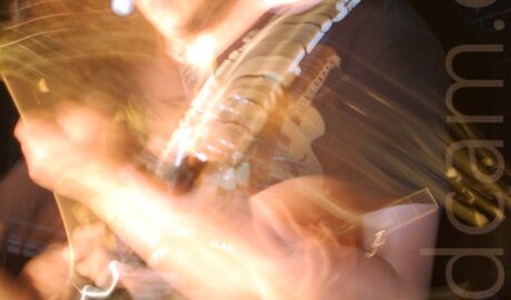 A long exposure shot of a man with long dark hair, wearing a black t-shirt and blue jeans, playing a black electric guitar. A along exposure and his movement have created several secondary images as he moves his head and body, as well as streaks of golden light. Black walls and ceiling fill the background.