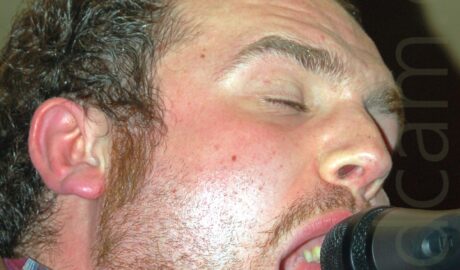 Close up of the head of a man facing to the right, singing into a microphone on a stand. The man has short dark air, and a stubbly beard, his mouth wide open and his eyes closed., with a brown leather jacket over a red and blue plaid shirt. Red walls and yellow ceiling fill the rest of the frame.