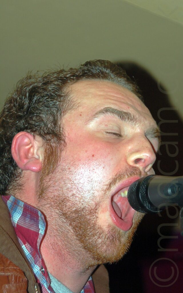 Close up of the head of a man facing to the right, singing into a microphone on a stand. The man has short dark air, and a stubbly beard, his mouth wide open and his eyes closed., with a brown leather jacket over a red and blue plaid shirt. Red walls and yellow ceiling fill the rest of the frame.