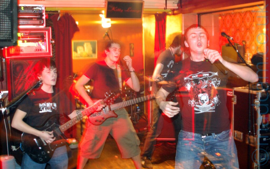 Four of five members of a band putting on a rather energetic performance on a stage. On the left is a fairly short young man with dark hair, wearing a black t-shirt with a white "The Idiot Rate" logo emblazoned across the chest, and playing a black electric guitar. In the middle is a man with short dark hair, wearing a plain black t-shirt and a beige pair of knee-length shorts, playing a brown electric guitar, with a look of surprise on his face. On the right is the bands singer, with short dark hair, wearing a black t-shirt with a white and red logo featuring a skull, dragons, and a pegasus, on the front. He is holding a microphone in his left hand, and is conspicuously not singing into it right now, but that is all about to change. Behind him is a tall man with shaggy brown hair and a brown beard, wearing a grey t-shirt, and playing a bass guitar. Orange and brown walls almost look golden in the yellow room lights, with red curtains hanging loose on some of the walls. Stacks of amps and monitors line some of the walls behind the band. A long exposure combines with the bands movement to create streaks of red light around them, along with secondary images through which the background can be seen.