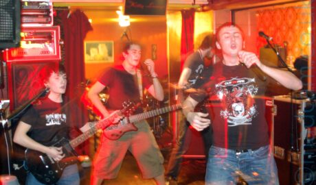 Four of five members of a band putting on a rather energetic performance on a stage. On the left is a fairly short young man with dark hair, wearing a black t-shirt with a white "The Idiot Rate" logo emblazoned across the chest, and playing a black electric guitar. In the middle is a man with short dark hair, wearing a plain black t-shirt and a beige pair of knee-length shorts, playing a brown electric guitar, with a look of surprise on his face. On the right is the bands singer, with short dark hair, wearing a black t-shirt with a white and red logo featuring a skull, dragons, and a pegasus, on the front. He is holding a microphone in his left hand, and is conspicuously not singing into it right now, but that is all about to change. Behind him is a tall man with shaggy brown hair and a brown beard, wearing a grey t-shirt, and playing a bass guitar. Orange and brown walls almost look golden in the yellow room lights, with red curtains hanging loose on some of the walls. Stacks of amps and monitors line some of the walls behind the band. A long exposure combines with the bands movement to create streaks of red light around them, along with secondary images through which the background can be seen.