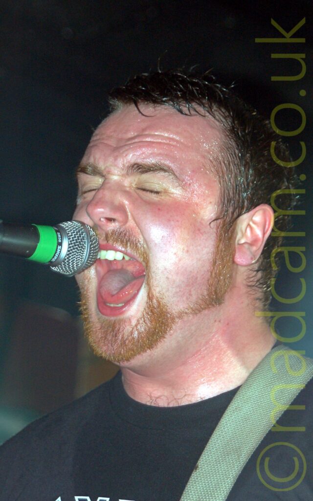 Close-up, head and shoulders shot of a man with short dark hair and a ginger goatee beard and sideburns, wearing a black t-shirt. He is singing in to a microphone on a stand in front of him. Stage lights behind him are casting a slight green halo around the back of his head.