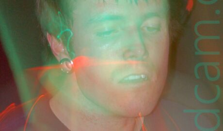 Close-up of the head and shoulders of a man with short dark hair, metal tunnels through his earlobes, and wearing a black t-shirt. A long exposure and a snap zoom have combined with green and red stage lights to create a green aura around him, and orange and red streaks of light around his head and chest.