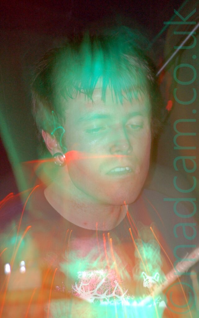 Close-up of the head and shoulders of a man with short dark hair, metal tunnels through his earlobes, and wearing a black t-shirt. A long exposure and a snap zoom have combined with green and red stage lights to create a green aura around him, and orange and red streaks of light around his head and chest.
