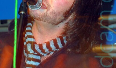 A man with mid-length dark hair and stubble along his chin line, wearing a dark blue denim shirt over a black and grey scarf, singing into a microphone on a stand, his eyes closed and head tilted to his left, looking almost blissed out. A long exposure and a snap zoom out has created yellow and orange light trails on the bottom half of the frame, with red and orange auras around his body. Blue walls and white ceiling fill the background at the top of the frame.