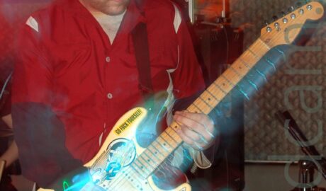 A man with short dark hair wearing a red shirt with grey shoulders, over a grey t-shirt, playing a yellow electric guitar that has been covered in multi-coloured stickers. A long exposure and his movement, combined with green, orange, and blue stage lights, create coloured secondary images and auras around him. Black and grey amplifiers and speakers line the black wall behind him.