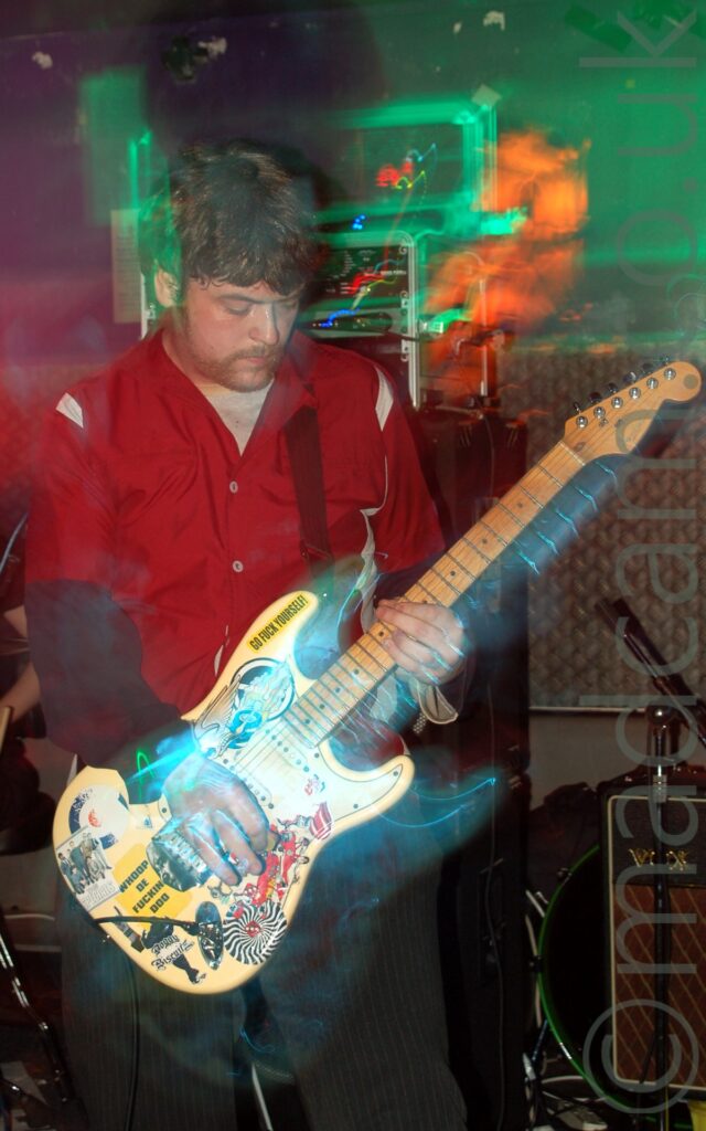 A man with short dark hair wearing a red shirt with grey shoulders, over a grey t-shirt, playing a yellow electric guitar that has been covered in multi-coloured stickers. A long exposure and his movement, combined with green, orange, and blue stage lights, create coloured secondary images and auras around him. Black and grey amplifiers and speakers line the black wall behind him.