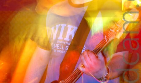 A man with floppy blond hair, wearing a blue t-shirt with black sleeves and black piping around the next, and white text "Nike" across the chest, as well as a blue pair of jeans, and playing a black and white bass guitar. A long exposure has combined with the stage lights and his movement to create golden secondary and tertiary images to his left and right, , as well as streaks of golden light. Purple walls, partially tinted red and orange, fill the background.