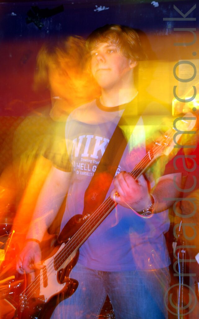 A man with floppy blond hair, wearing a blue t-shirt with black sleeves and black piping around the next, and white text "Nike" across the chest, as well as a blue pair of jeans, and playing a black and white bass guitar. A long exposure has combined with the stage lights and his movement to create golden secondary and tertiary images to his left and right, , as well as streaks of golden light. Purple walls, partially tinted red and orange, fill the background.