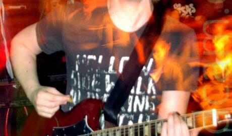 A man with floppy, shoulder-length blond hair, wearing a black t-shirt with faded white text filling the front, and a blue pair of jeans, playing a red electric guitar. An orange pole stretches from floor to ceiling behind him, painted to look sticky and organic and visceral, with some people beyond that, with dark coloured walls behind them. A long exposure and his movement have created red and orange streaks of light and secondary images.